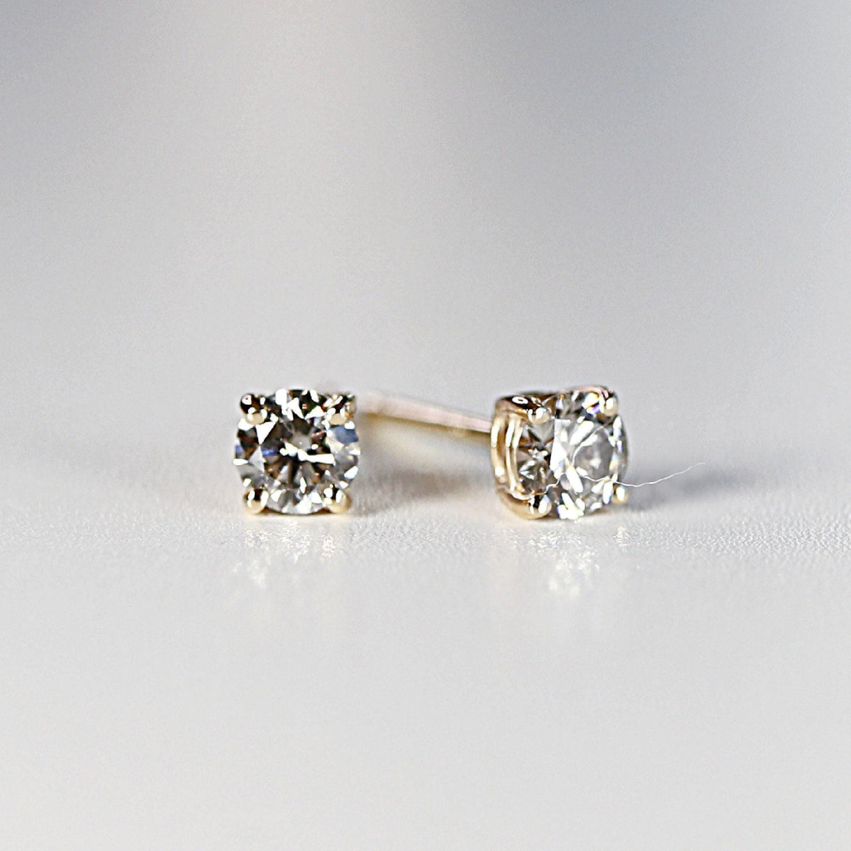 Two round brilliant-cut diamond stud earrings in yellow gold settings displayed on a light surface.