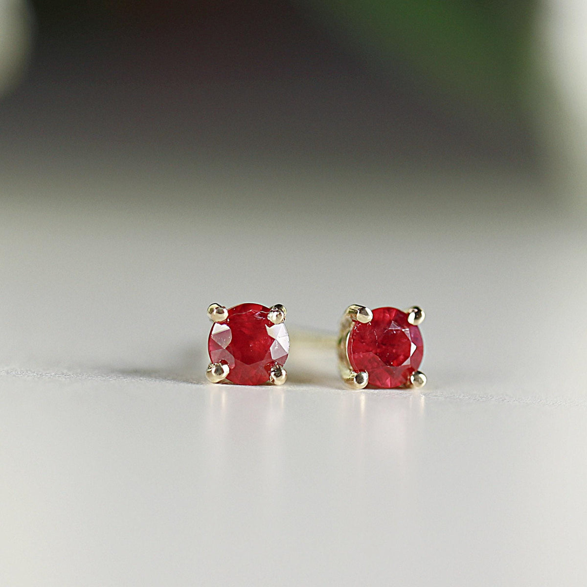 Two round red ruby stud earrings in four-prong gold settings standing upright on a light surface.