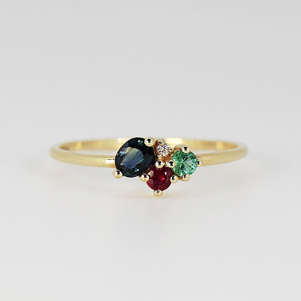 A thin gold ring with a cluster of four gemstones including an oval blue sapphire, red ruby, green emerald, and a tiny white diamond set against a plain light background.