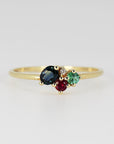 A thin gold ring with a cluster of four gemstones including an oval blue sapphire, red ruby, green emerald, and a tiny white diamond set against a plain light background.