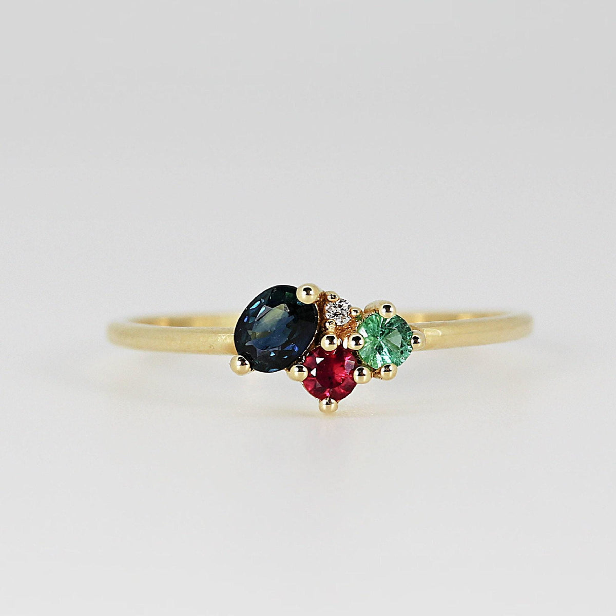 Direct macro shot of the gold ring centered horizontally, displaying the dark blue oval sapphire next to the round red and green stones and small accent diamond.