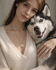 A woman wearing a cream V-neck sweater hugs a live husky while wearing the gold husky pendant necklace.