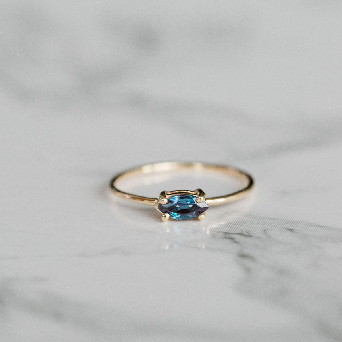 A gold ring with a horizontally set marquise-cut blue and purple gemstone resting on a white marble surface.