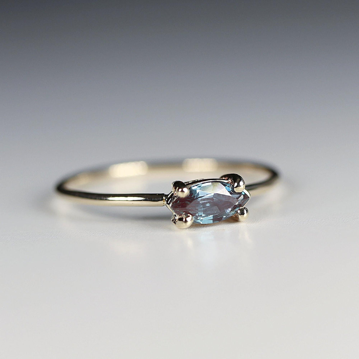 A close-up of a gold ring with a horizontal marquise-cut blue-purple gemstone secured by four prongs on a neutral surface.