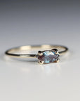 A close-up of a gold ring with a horizontal marquise-cut blue-purple gemstone secured by four prongs on a neutral surface.