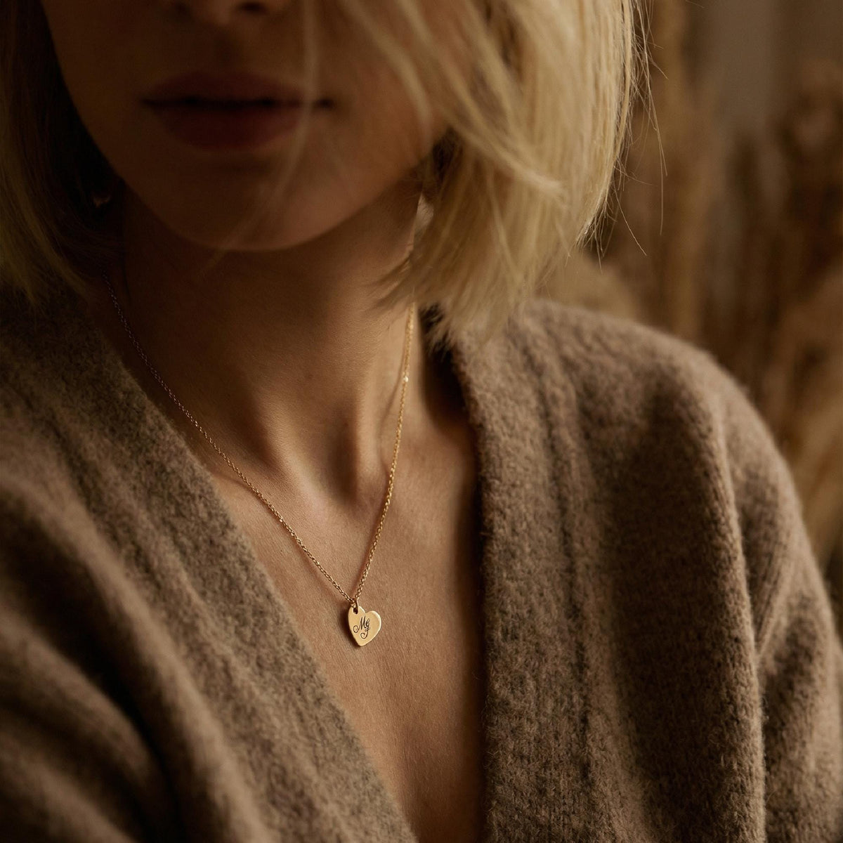 A person wearing a gold heart pendant necklace engraved with script initials against a dark knitted background.