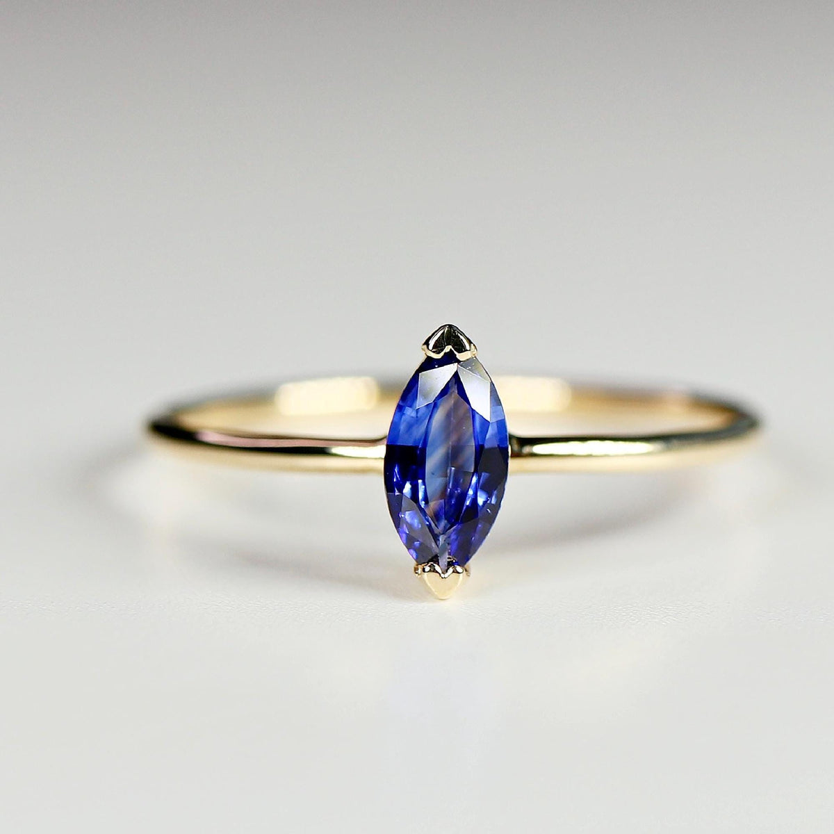 A thin gold ring featuring a prong-set marquise-cut blue sapphire solitaire, shown from the front on a light surface.