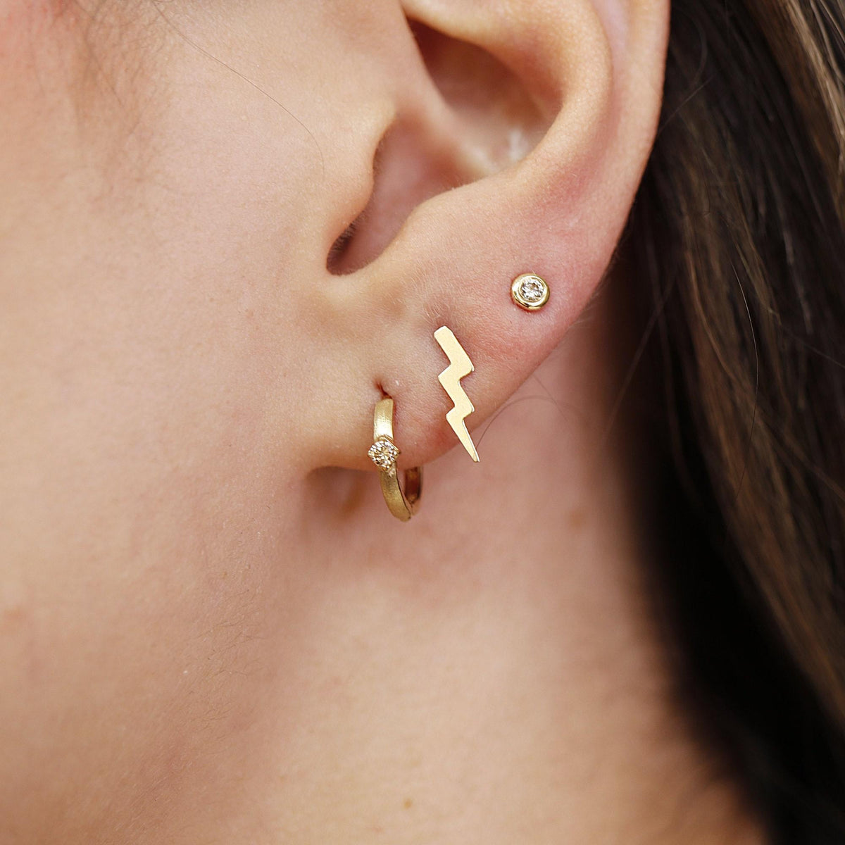 Side view of an earlobe showing a gold diamond huggie hoop earring alongside a lightning bolt stud and a bezel-set round stone stud.