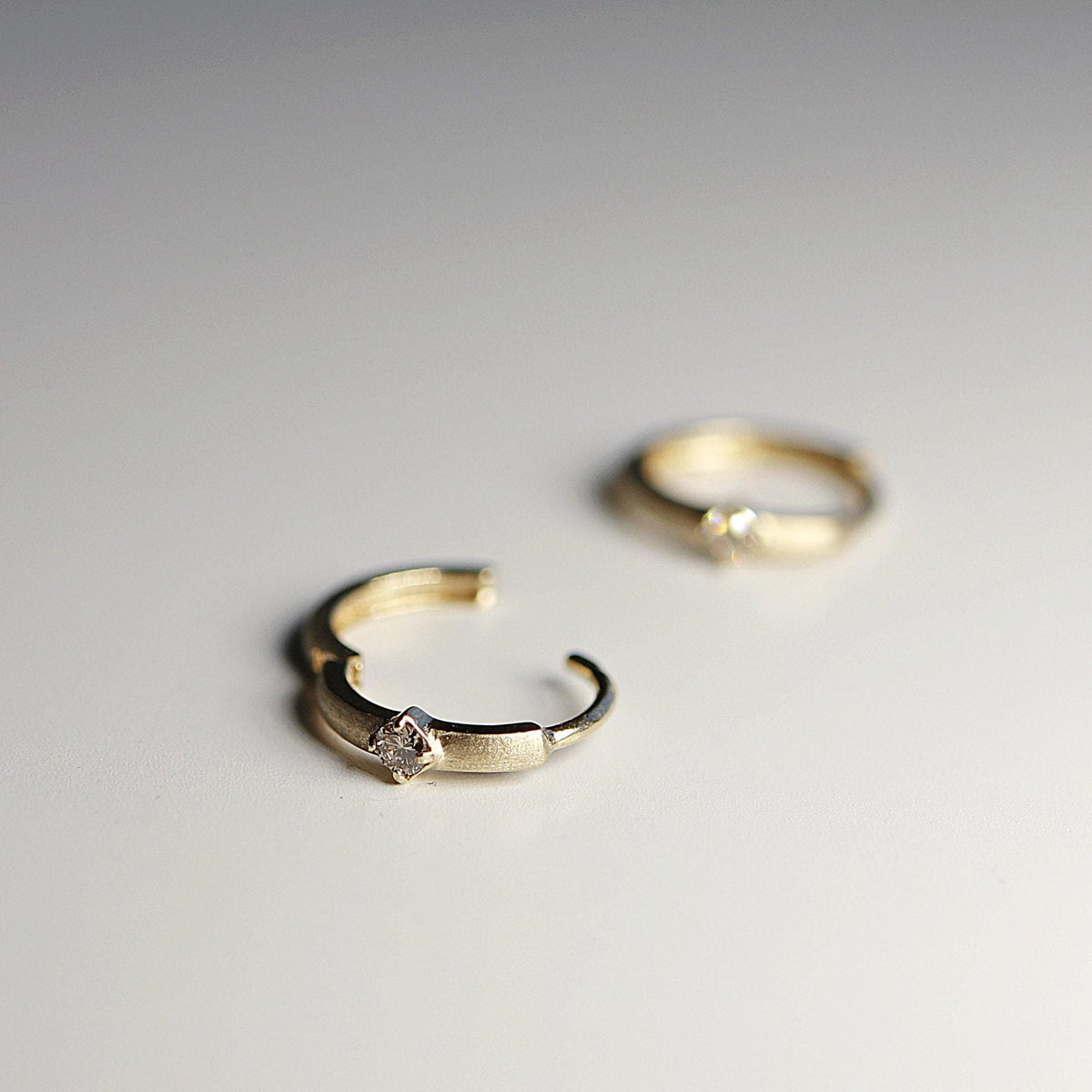 Two gold huggie hoop earrings sit on a white surface, with one hoop open in the foreground and the second hoop blurred in the background.