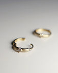 Two gold huggie hoop earrings sit on a white surface, with one hoop open in the foreground and the second hoop blurred in the background.