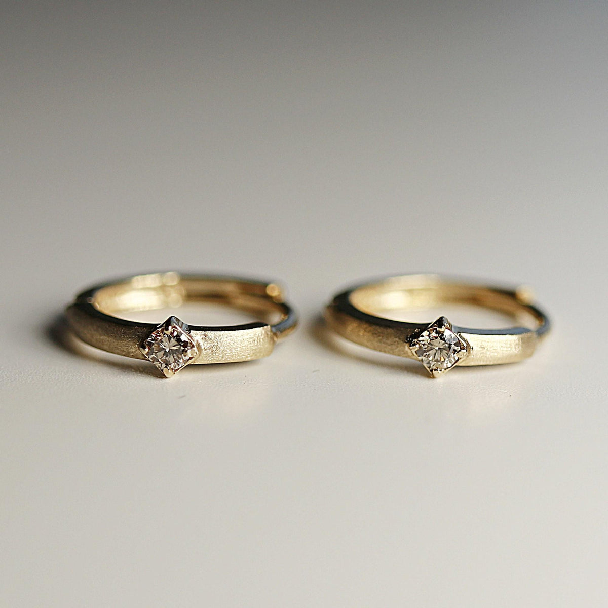 Two matte-finish gold hoop earrings with a single diamond set in a square prong mounting stand side by side on a flat white surface.