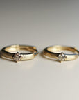 Two matte-finish gold hoop earrings with a single diamond set in a square prong mounting stand side by side on a flat white surface.
