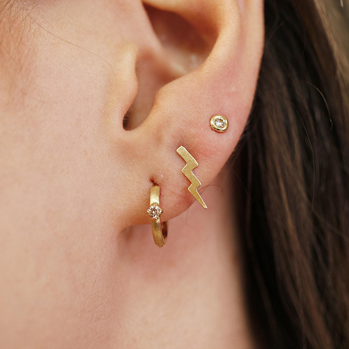 Close-up of a pierced ear wearing a gold diamond huggie hoop earring, a gold lightning bolt stud, and a round diamond stud.