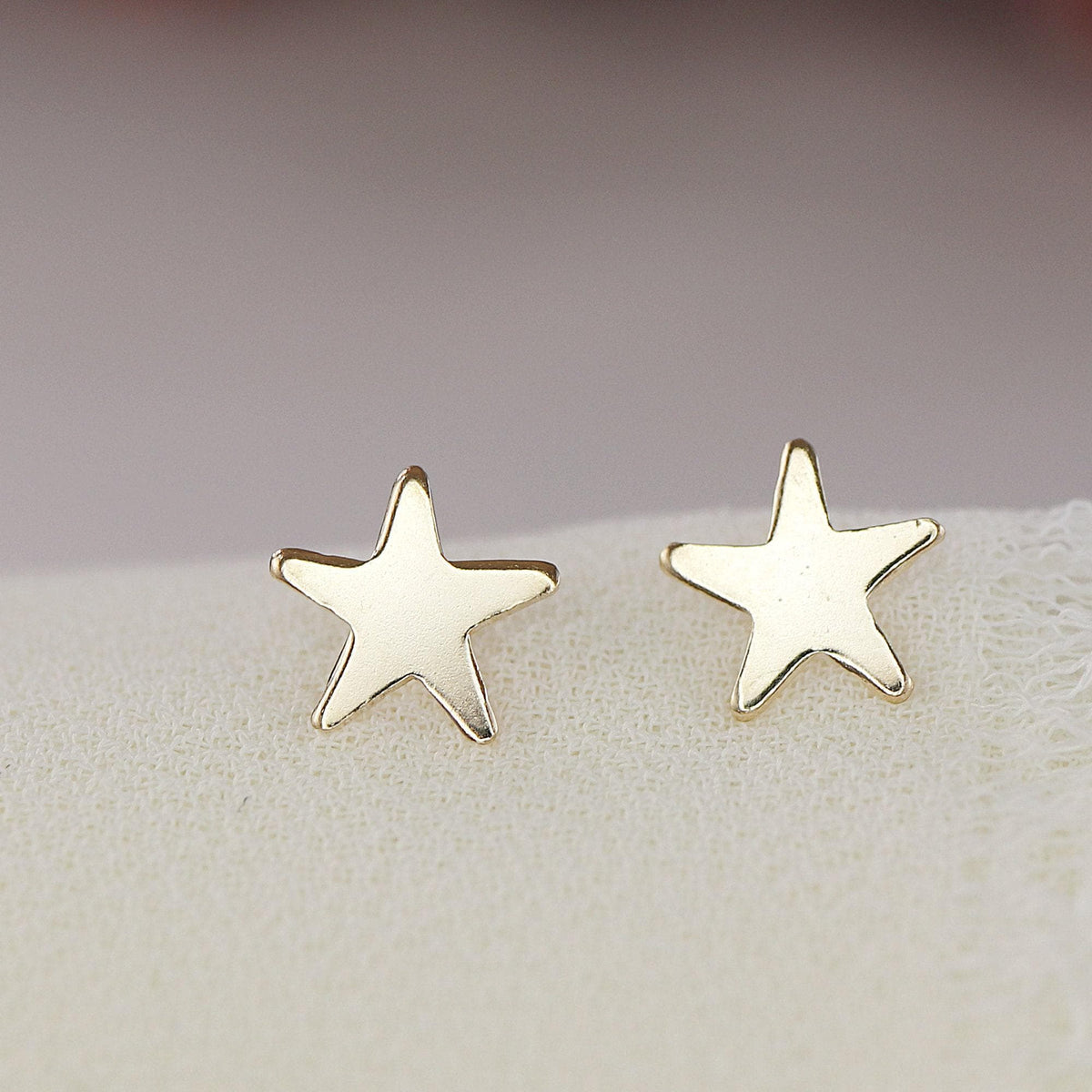 Two gold star stud earrings rest side by side on a soft white fabric surface.