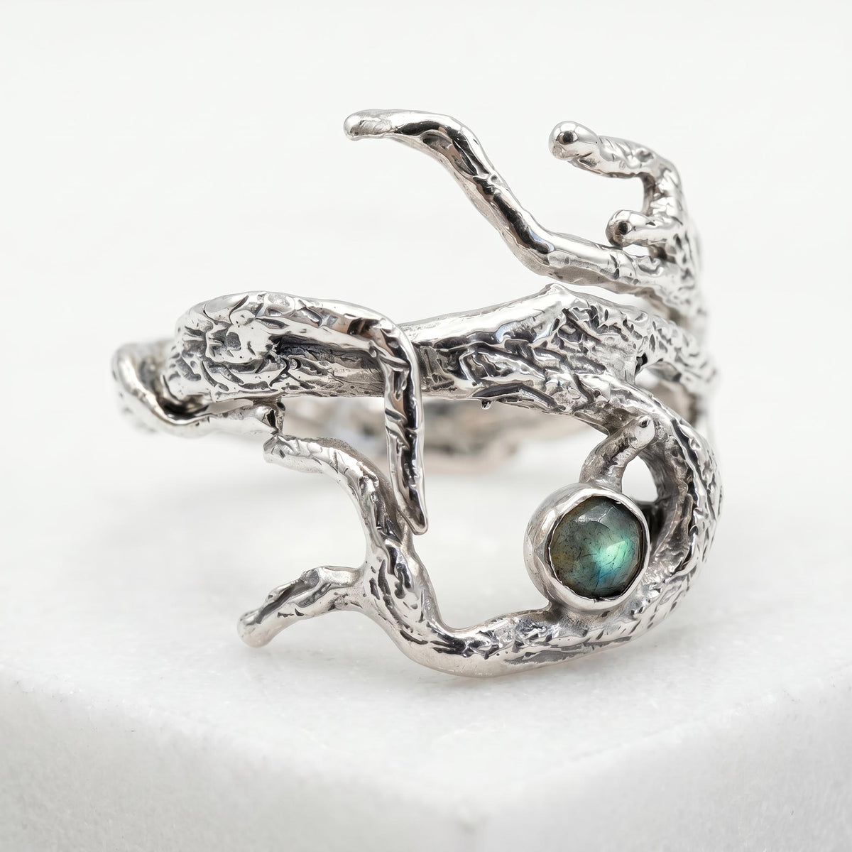 A silver ring designed like rough twigs and tree bark holding a round grey gemstone.