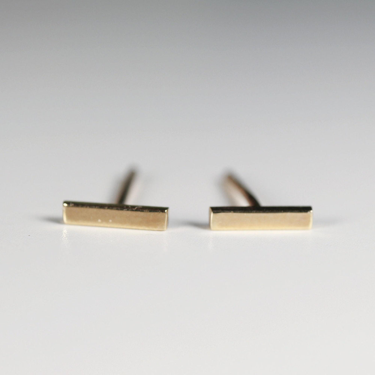 Two small gold bar stud earrings resting on a white surface with their post backs visible.
