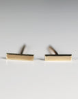 Two small gold bar stud earrings resting on a white surface with their post backs visible.
