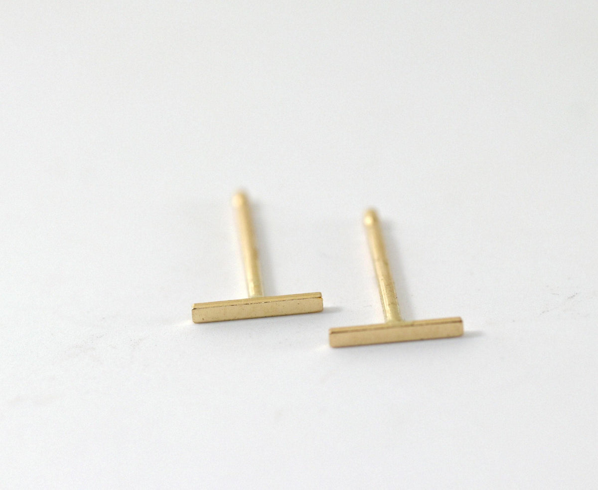 A pair of minimalist gold bar stud earrings standing upright on a light surface.