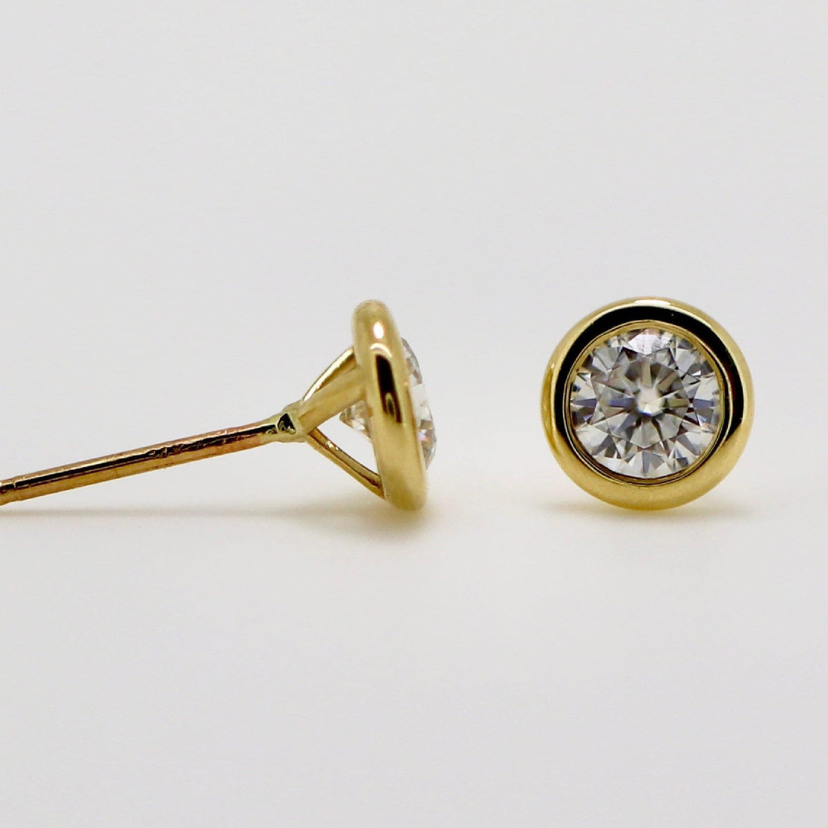 Two gold bezel-set stud earrings shown against a white background, with one earring facing forward and the other angled to show the post.