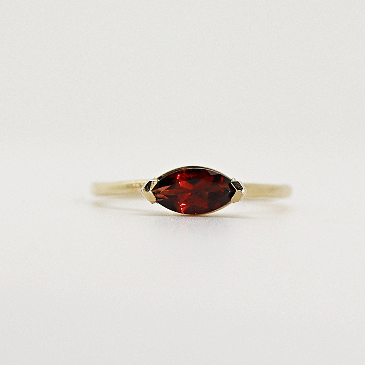 Studio shot of a gold ring centered horizontally with a dark red marquise-cut gemstone against a light background.