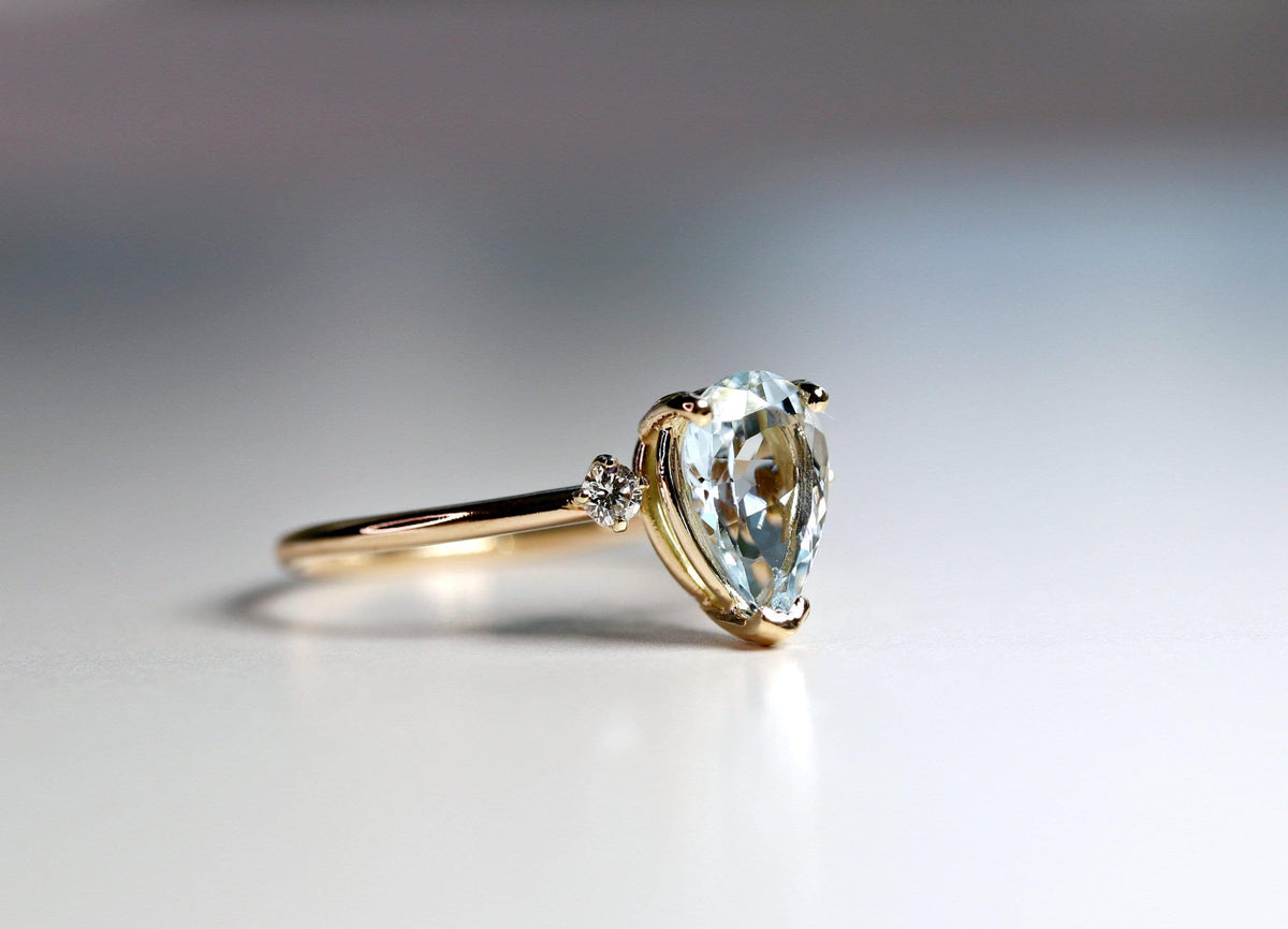 A gold ring featuring a pear-shaped light blue gemstone and a small round accent diamond on one side, shown in profile on a light surface.