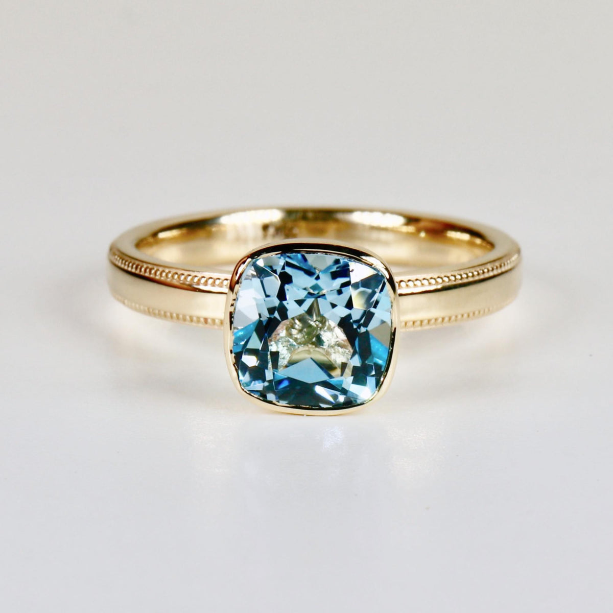 A front view of a gold ring featuring a bezel-set cushion-cut light blue gemstone with milgrain detailing along the band on a white surface.