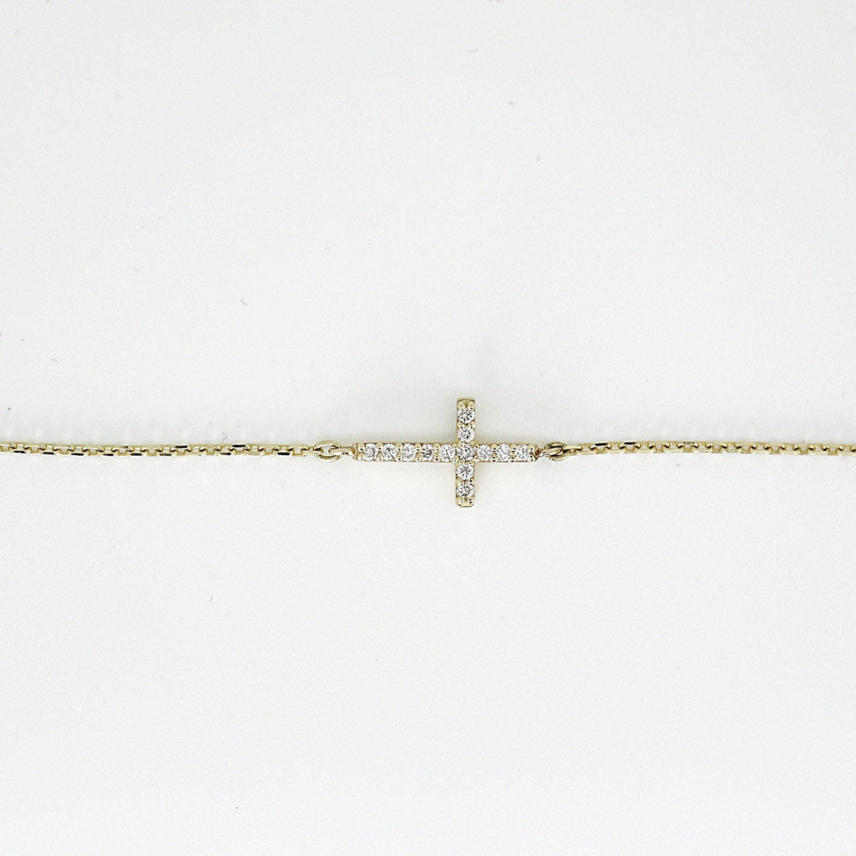 A close-up shot on a white background shows the central pavé diamond cross element on a thin gold chain bracelet.