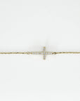 A close-up shot on a white background shows the central pavé diamond cross element on a thin gold chain bracelet.