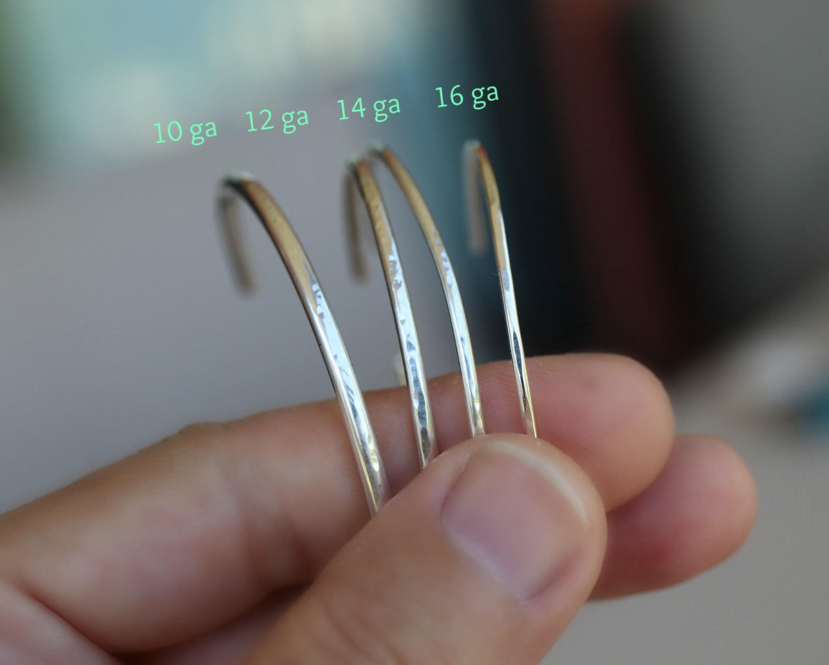 A hand holding four silver cuff bracelets of varying thickness, labeled with green text as 10 ga, 12 ga, 14 ga, and 16 ga.