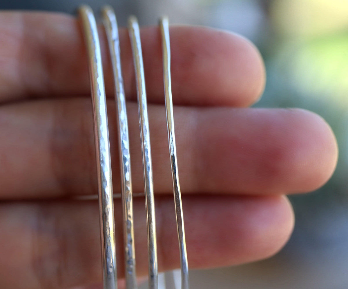 Close-up of four thin silver hammered cuff bracelets held vertically across a person fingers.