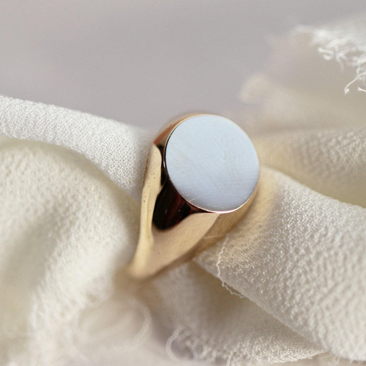 A smooth gold signet ring rests on a textured white fabric.