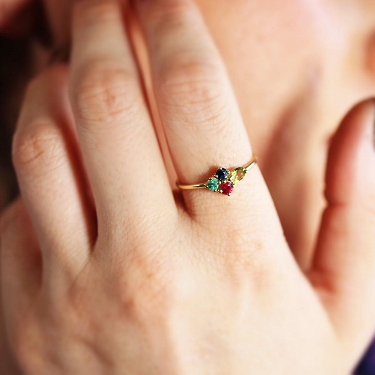 Close-up of a gold ring with multiple colorful gemstones worn on a finger near a person face.