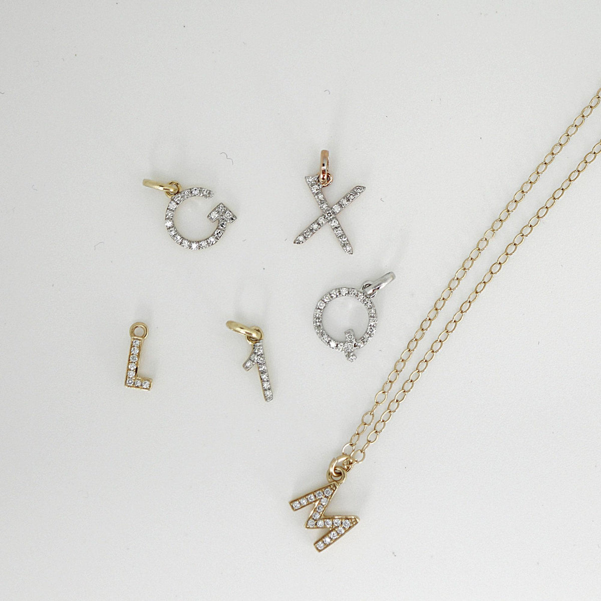 Several gold and silver letter and number charms with diamonds laid flat next to a gold chain necklace with a letter M pendant.