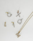 Several gold and silver letter and number charms with diamonds laid flat next to a gold chain necklace with a letter M pendant.