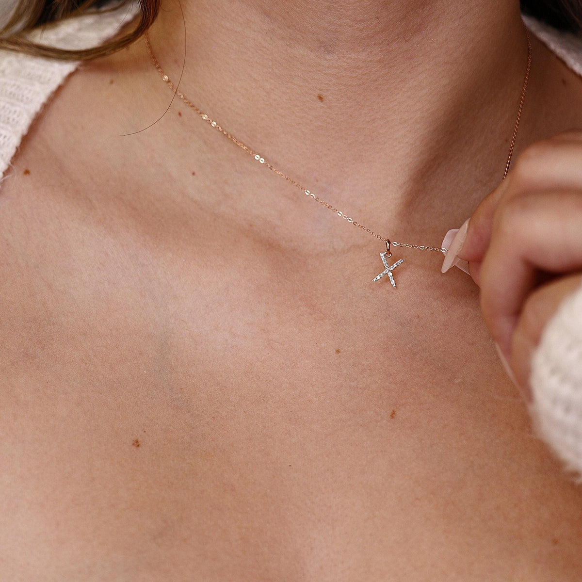 A person wearing a delicate gold chain necklace with a diamond-paved letter X pendant.