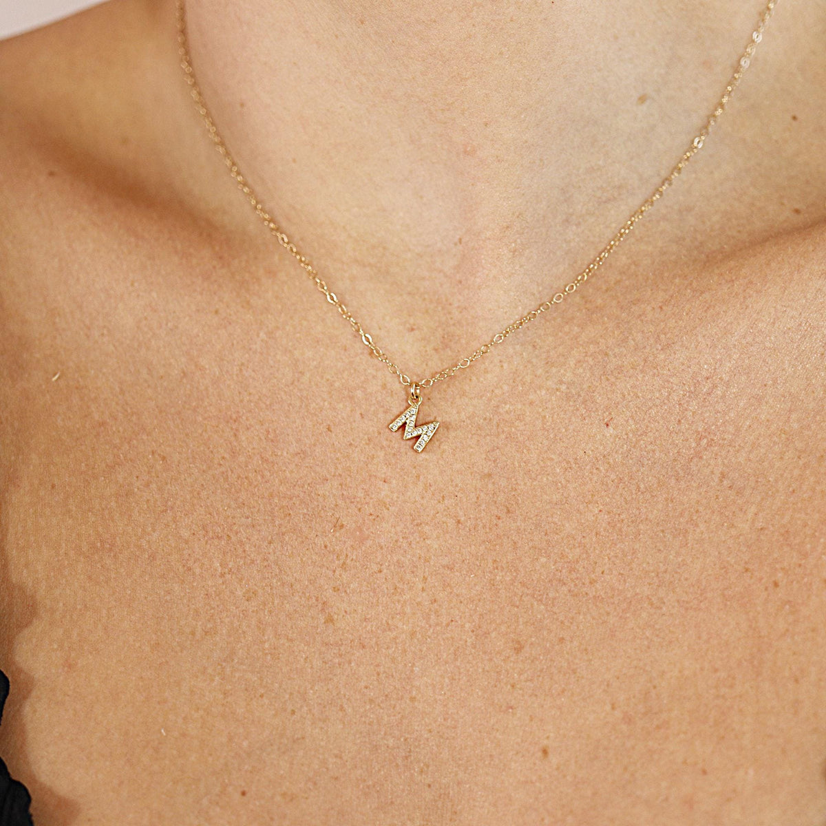 A close-up of a person wearing a dainty gold chain necklace with a small diamond letter M pendant.