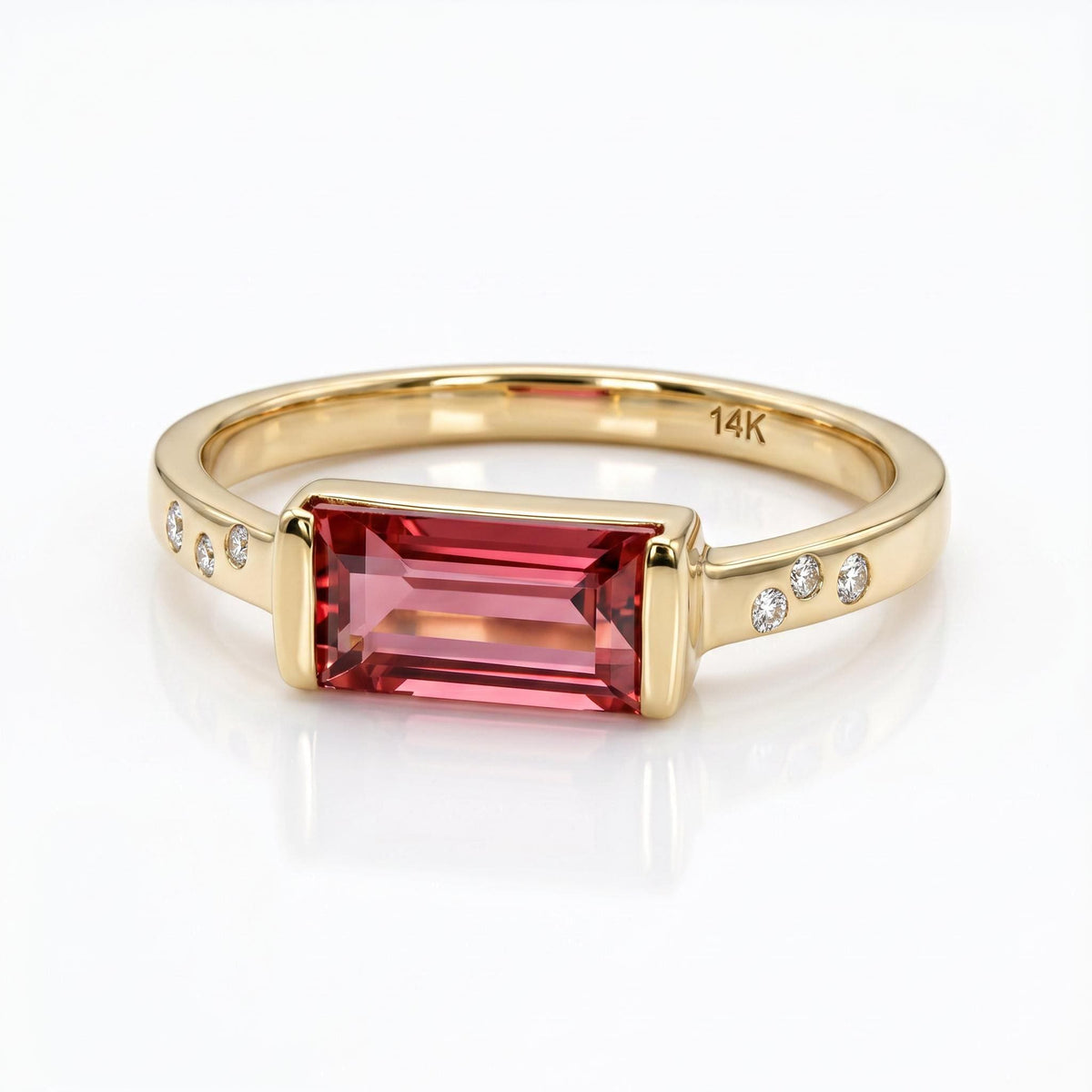 A gold ring featuring an east-west set rectangular pink tourmaline baguette and three small flush-set round diamonds on each shoulder.