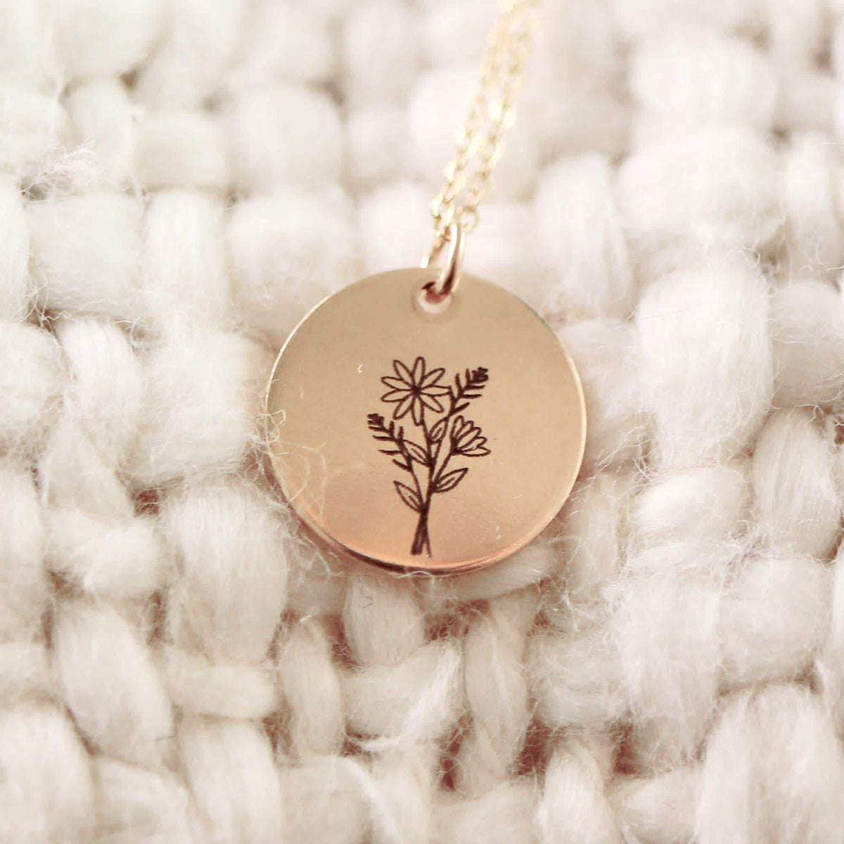 A close-up of a gold disc necklace pendant engraved with a floral bouquet resting on a white textured knit fabric.