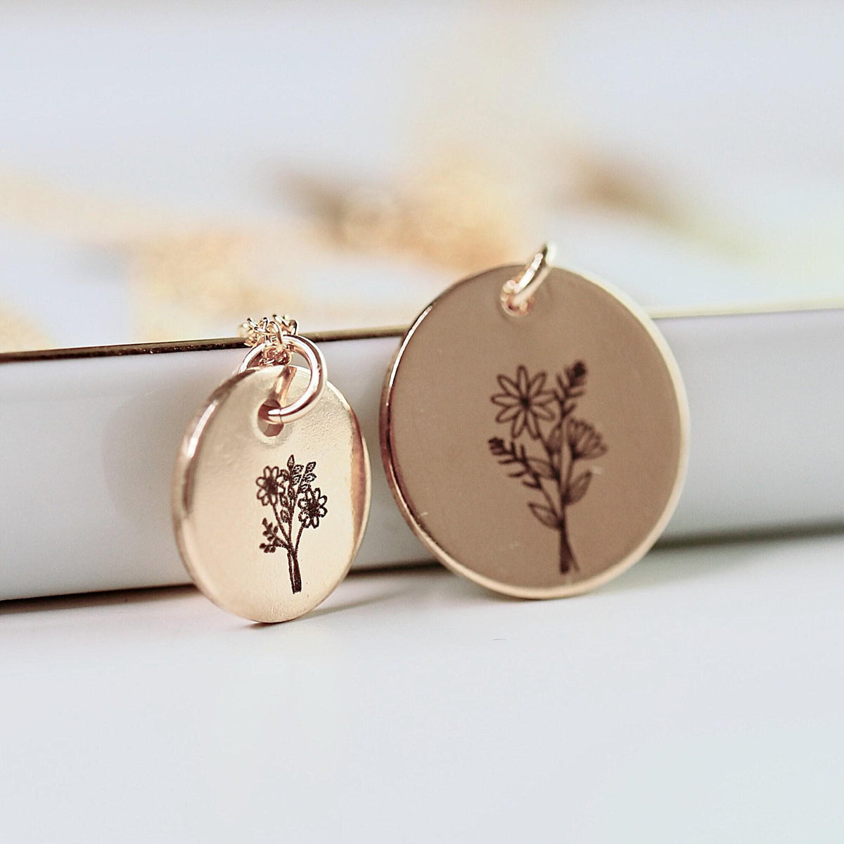 Two rose gold disc pendants of different sizes with engraved flower designs, displayed in front of a white surface.