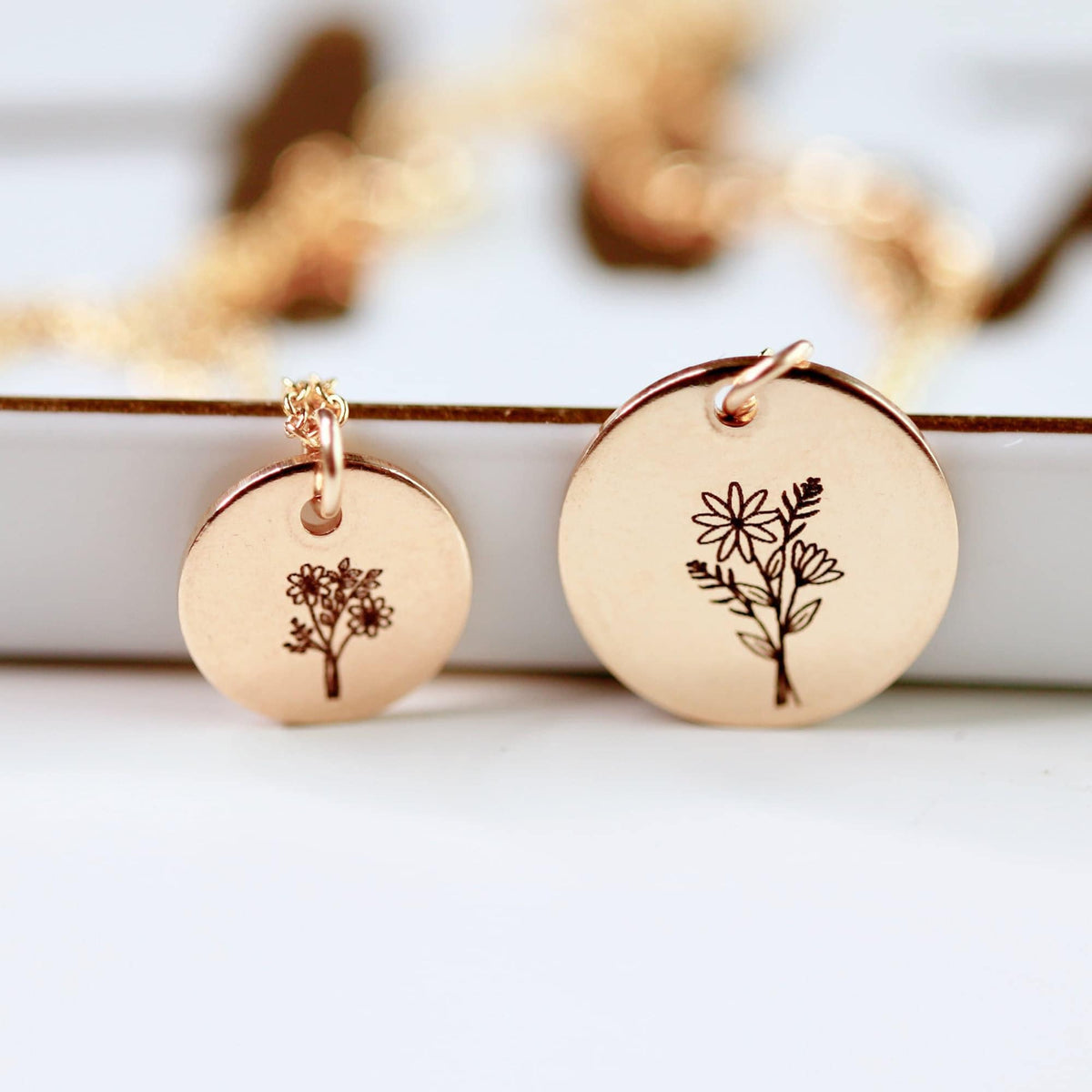 Two rose gold round pendants engraved with floral arrangements, hanging from chains against a white background.