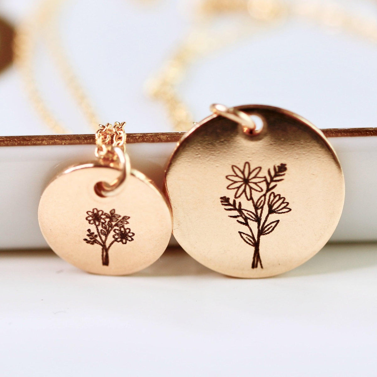 Two gold-colored engraved disc pendants with floral illustrations shown close up against a white ledge.