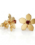 Two gold flower stud earrings featuring faceted dark red gemstone centers on a white surface.