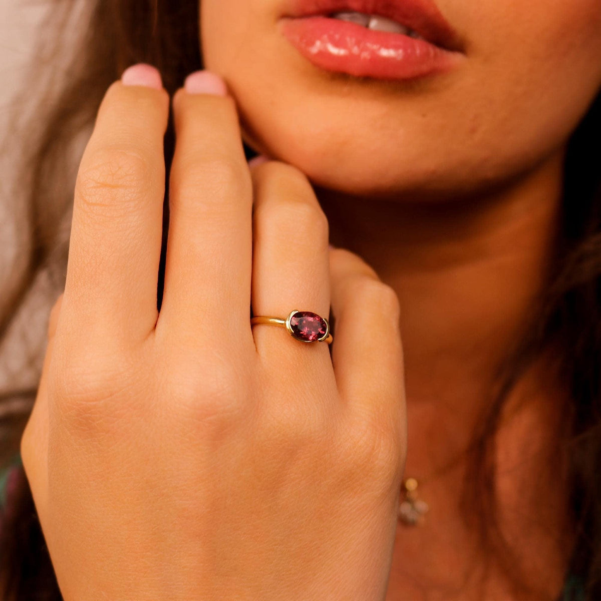 A close-up shot of a person wearing a gold ring featuring a horizontally set oval pink tourmaline stone on their finger.
