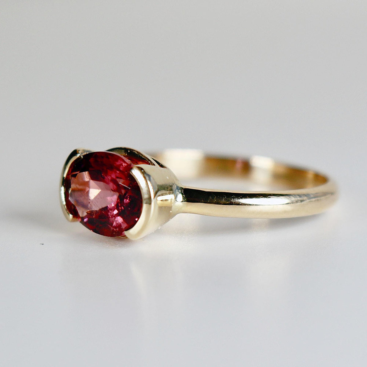 A side view of a gold ring with an east-west set oval pink tourmaline gemstone in a half-bezel setting against a plain light background.
