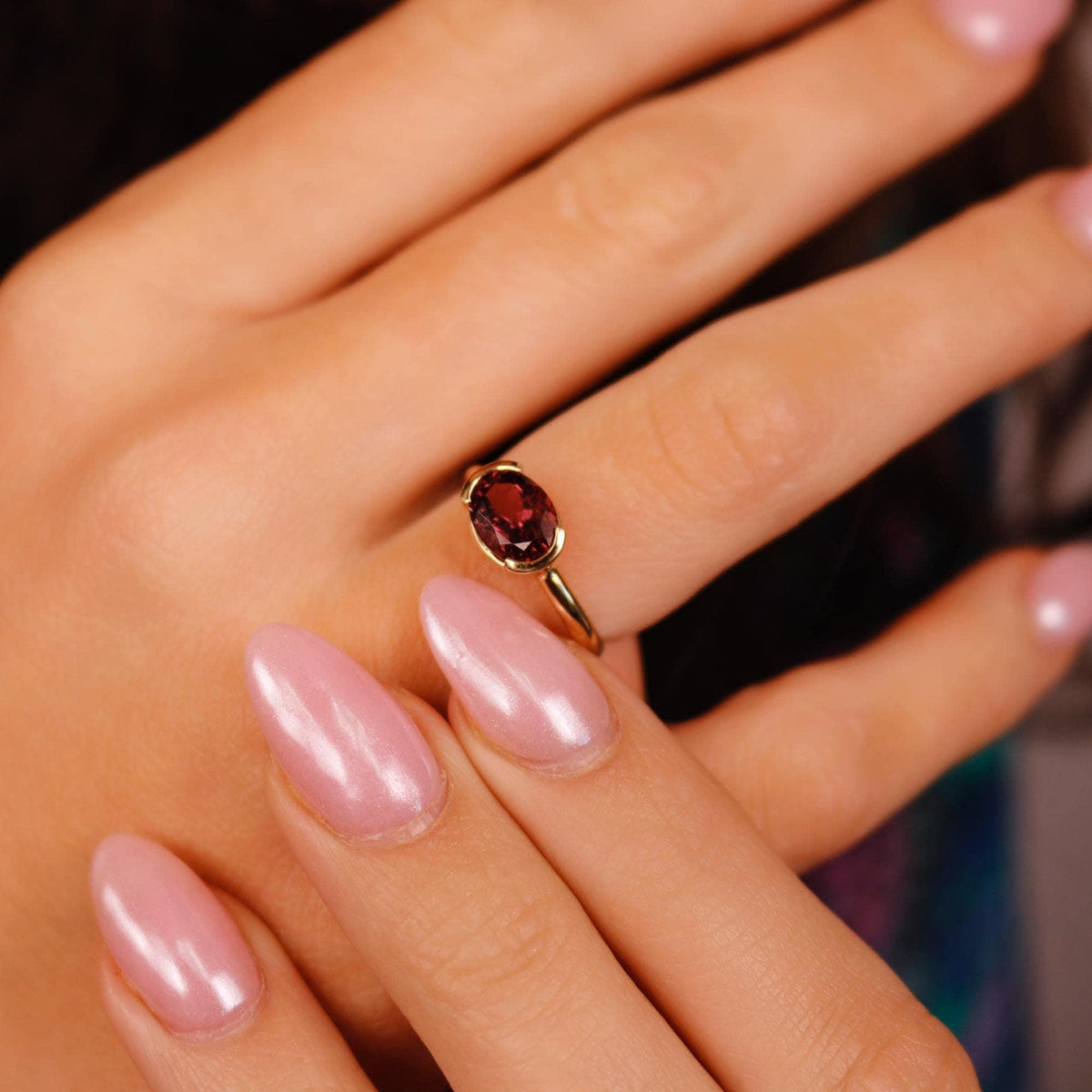 A close-up view of a hand with glossy light pink nails wearing a gold ring with a vertical oval pink tourmaline stone.