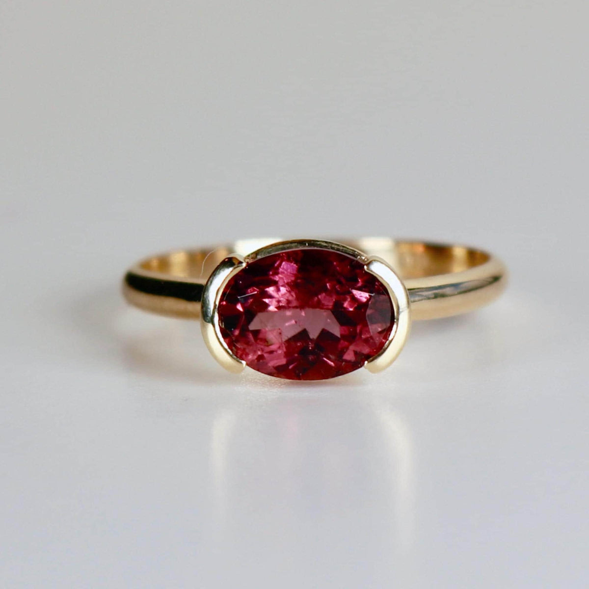 A straight front view of the gold ring centered on a white surface, highlighting the deep pink oval tourmaline gemstone.