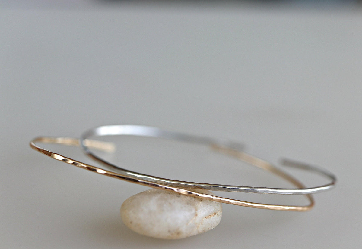 Two thin metal open cuff bracelets balance on top of a smooth light-colored stone against a plain light background.