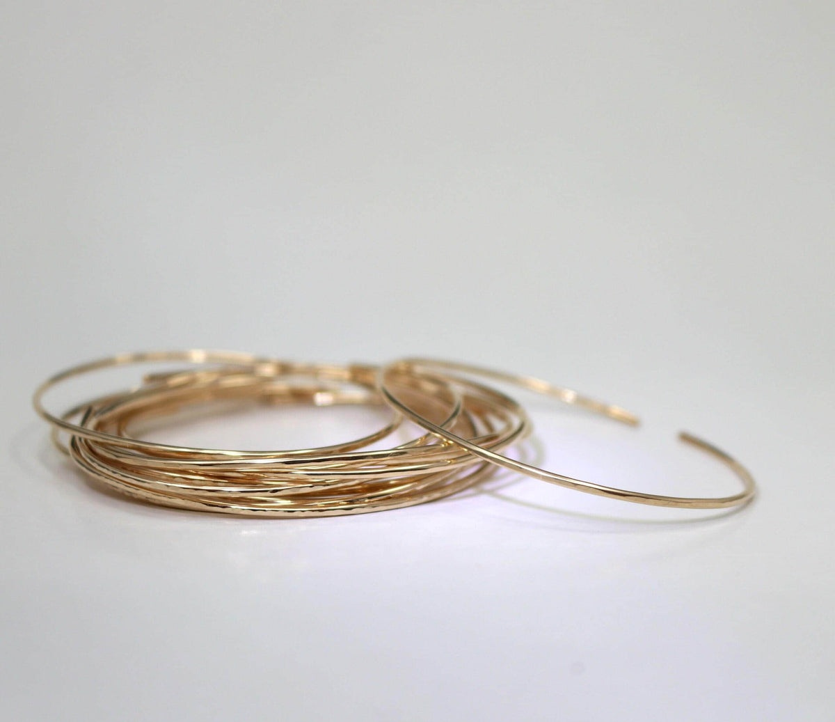A large stack of thin yellow gold hammered cuff bracelets is arranged on a smooth white surface.