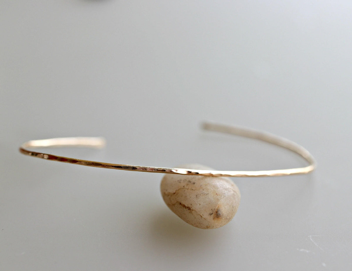 A single thin hammered gold cuff bracelet is propped up by a smooth round stone against a light background.