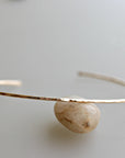 A single thin hammered gold cuff bracelet is propped up by a smooth round stone against a light background.